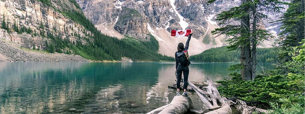 women hiking along a river in the mountains standing on a log holding a Canadian flag