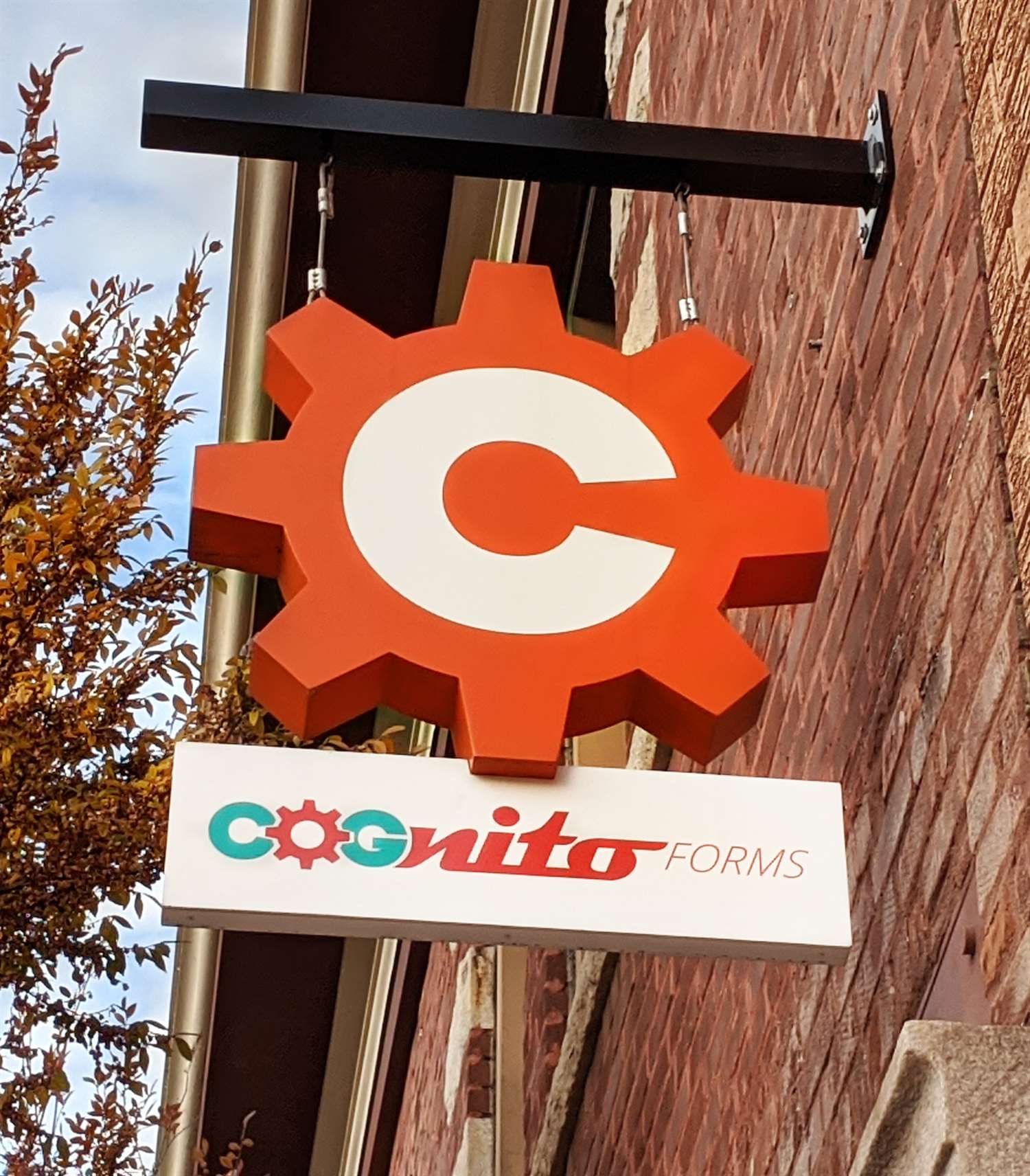 A Cognito Forms outdoor logo sign, featuring an orange gear and white "C" logo, mounted on a brick wall.