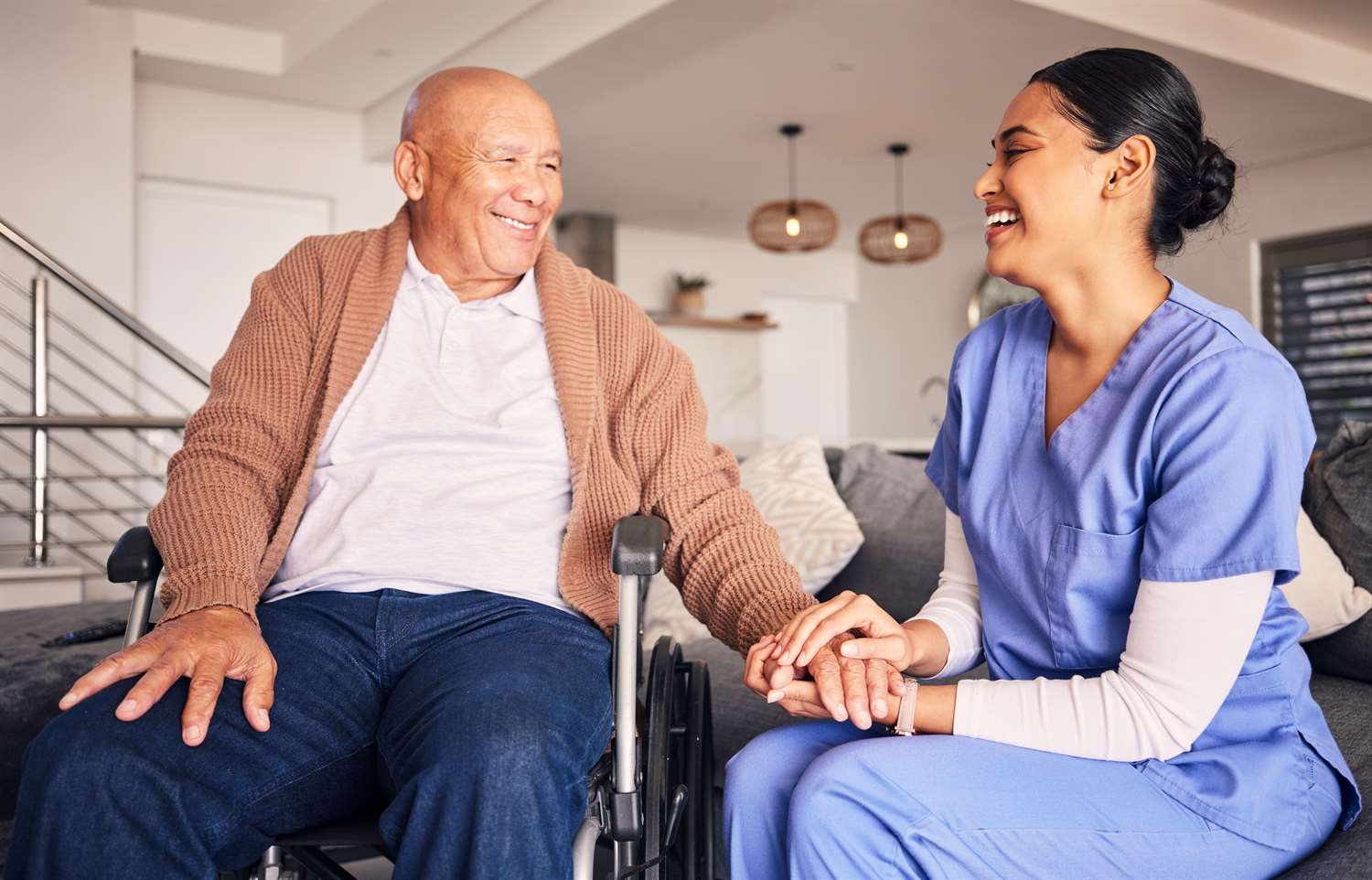 A nurse in a blue uniform smiling and holding the hand of a client in a wheelchair.