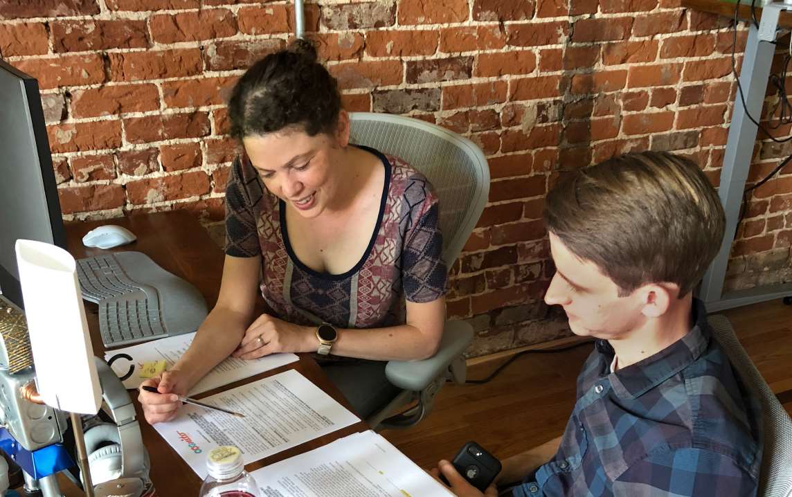 Two people collaborating over documents at a desk in a brick-walled office.